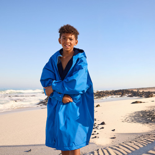 A young boy wearing a large blue jacket stands on a sunny beach. The ocean waves crash behind him, creating a serene and lively atmosphere.