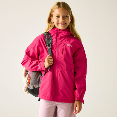 A young girl smiles, wearing a vibrant pink jacket and holding a black backpack. The background is neutral, creating a cheerful, outdoor-ready vibe.