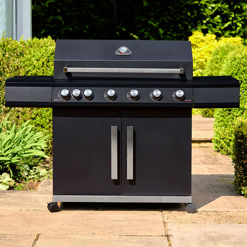 Black six-burner barbecue with closed hood, wide side shelves and double-door cupboard, shown on a garden patio.
