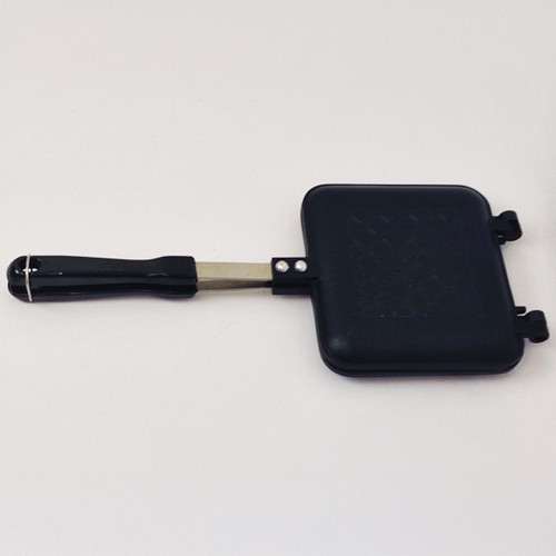 A black square sandwich press with a handle, designed for grilling or toasting sandwiches, rests on a light surface.