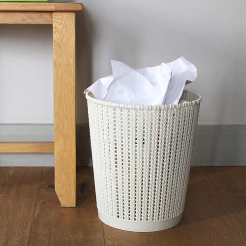 A cream waste paper bin under a desk with some paper sticking out