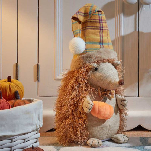 A plush hedgehog toy wearing a plaid hat, holding a small orange pumpkin, with a backdrop of decorative pumpkins.
