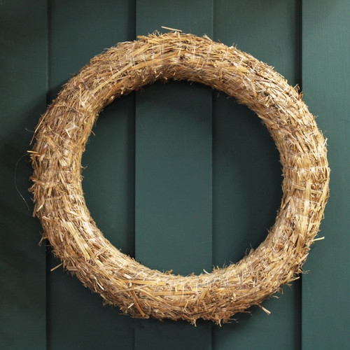 A circular natural straw wreath base made from tightly bundled golden straw, secured with fine white thread, hanging against a dark green wooden door.
