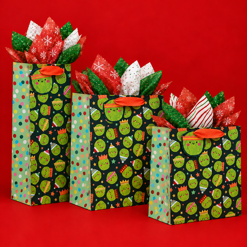 Three Christmas gift bags in different sizes, featuring playful green Brussels sprouts wearing festive hats and crowns on a dark background, with colourful polka-dot side panels and orange ribbon handles. Filled with red, green, and white patterned tissue paper, arranged against a bright red background.