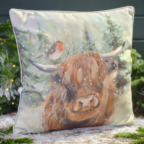 A decorative pillow featuring a Highland cow with snowy fur, a robin perched nearby, and a snowy forest background.