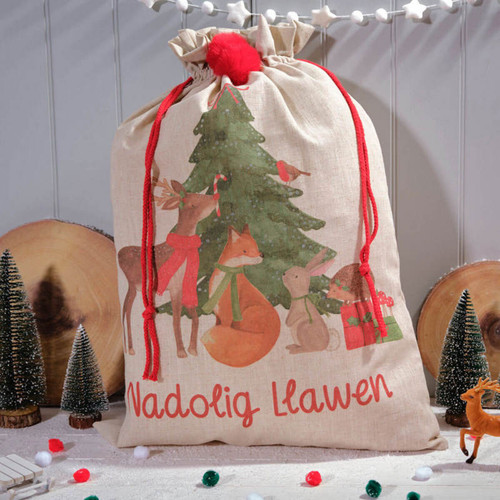 A festive burlap gift bag featuring woodland animals around a Christmas tree, with the words "Nadolig Llawen." Decorated with red cords and pom.