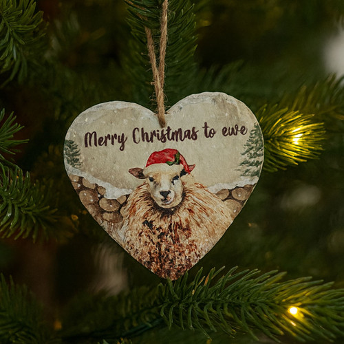 A heart-shaped Christmas ornament featuring a sheep in a red Santa hat and the words 'Merry Christmas to ewe' hangs on a decorated evergreen tree, surrounded by warm white fairy lights and lush green pine needles.