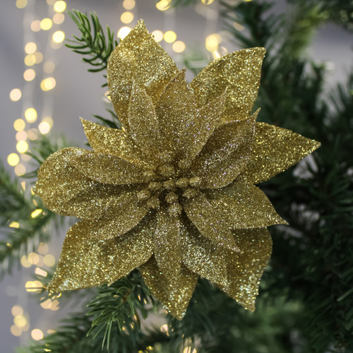 A sparkling gold poinsettia ornament decorates a lush green Christmas tree, with soft, warm fairy lights blurred in the background, creating a festive ambiance.
