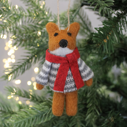 Felt fox ornament with red scarf and striped jumper hangs on a Christmas tree. Soft lighting and evergreen branches evoke a cosy, festive mood.