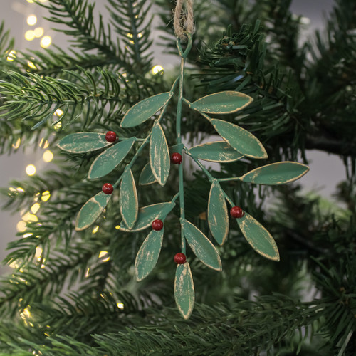 A rustic, metal mistletoe ornament hangs on a Christmas tree, adorned with green leaves and red berries. Soft, warm lights create a festive, cozy ambiance.