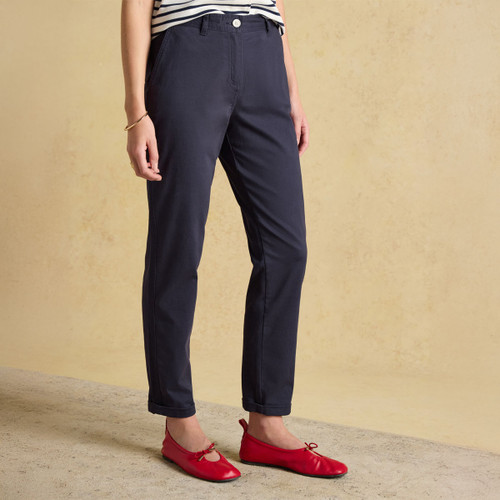 A woman stands in a relaxed pose wearing navy chino trousers and bright red shoes, set against a textured beige background.