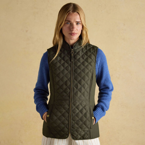 A person stands against a beige background, wearing a quilted olive green gilet over a blue sweater, showcasing a stylish layered outfit.