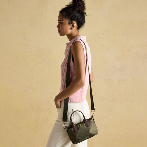A woman in a pink sleeveless sweatshirt and white trousers holds an khaki green handbag, posed against a soft beige background.
