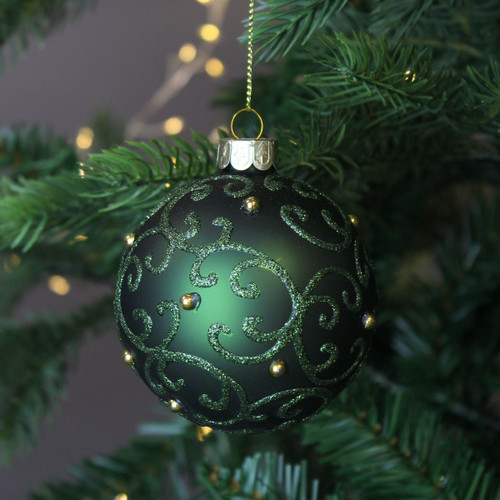 A view of a green glass bauble with a glittery green filigree pattern hanging on a Christmas tree