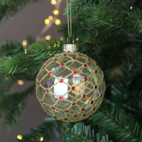 A view of a clear glass bauble with a glittery gold trellis design with red dots hanging on a Christmas tree