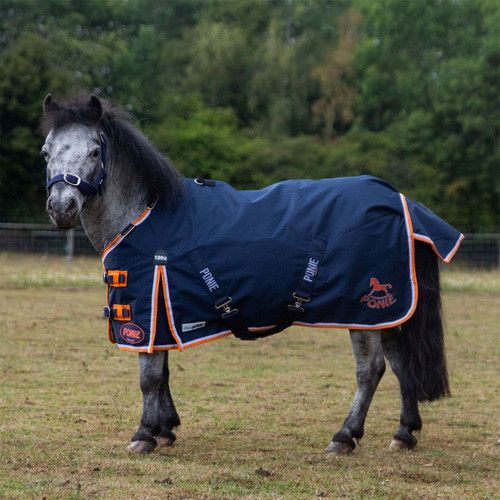 A small grey pony stands in a grassy paddock wearing a navy turnout rug with bright orange and white trim, a navy halter, and visible “PONIE” branding.