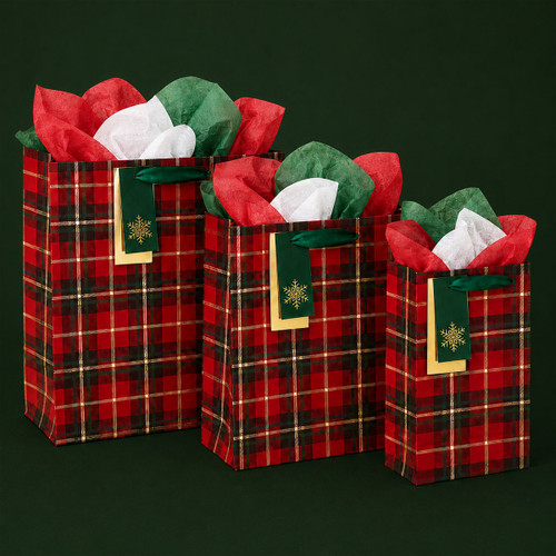 Three red tartan Christmas gift bags in different sizes with green ribbon handles, snowflake gift tags and red, green and white tissue paper.
