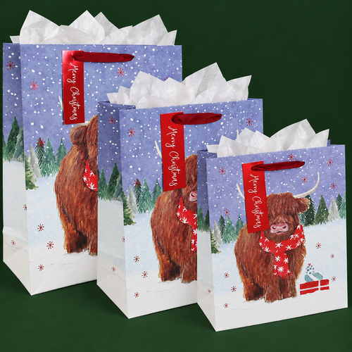 Three Christmas gift bags in different sizes, filled with white tissue paper and arranged against a dark green background. Each features a shaggy Highland cow wearing a red snowflake scarf in a snowy forest, with red ribbon handles and matching “Merry Christmas” gift tags.