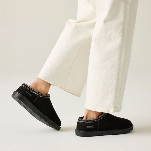 Black suede slip-on shoes with decorative braided trim and textured black soles, worn with white wide-leg trousers.