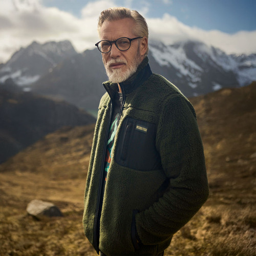 A bearded man wearing glasses and a green fleece jacket stands confidently against a backdrop of majestic snow-capped mountains under a partly cloudy sky.
