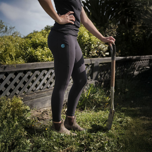 A person stands confidently beside a spade in a garden, wearing dark leggings and boots. Sunlit greenery and a lattice fence are in the background.