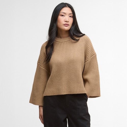 Woman with long hair stands against a plain background wearing a loose, camel-coloured knit sweater and black pants, exuding a relaxed, casual vibe.