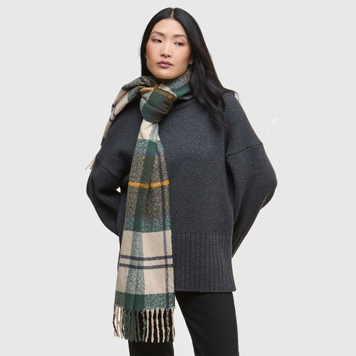 Woman in grey sweater and checked scarf stands against a plain background. She looks contemplative, arms relaxed. Cosy, casual fashion vibe.