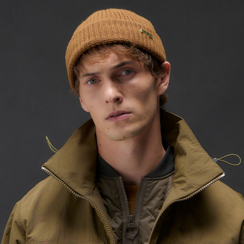 A young man wearing a brown knit beanie and olive jacket with a zipper, gazing seriously at the camera. Dark, neutral background. Cosy, casual vibe.