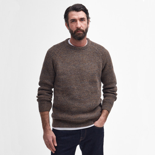 Man with a beard wearing a textured brown sweater and dark jeans, standing against a plain white background, hands in pockets, exuding a relaxed vibe.