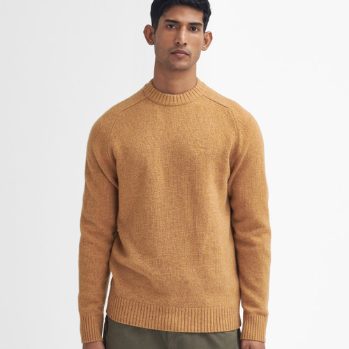 A person wearing a warm, light brown sweater with ribbed cuffs and neckline stands against a plain white background, exuding a calm, composed demeanour.