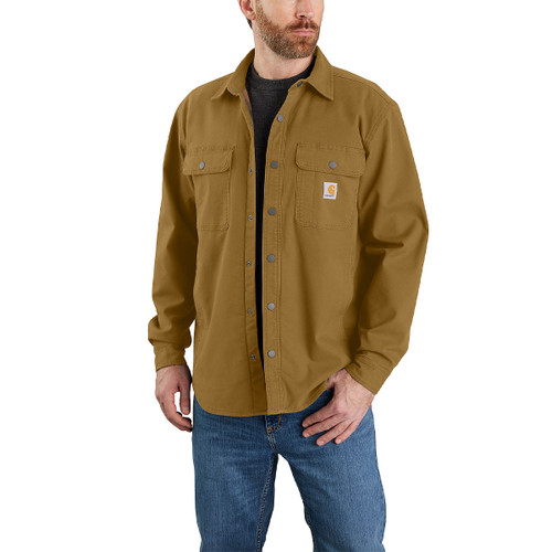 Man wearing a Carhartt tan button-up work shirt with dual chest flap pockets, left pocket featuring the Carhartt logo patch, styled with a black T-shirt and blue jeans, against a white background.