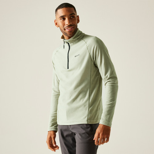 A man in a light green zip-up fleece and dark pants stands against a beige background, smiling softly. Casual, relaxed, and stylish athletic look.