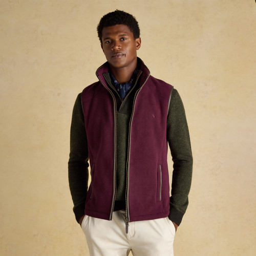 Man wearing a burgundy-coloured fleece gilet with a high collar and zip front, layered over a dark green jumper and navy shirt with white polka dots. He is also wearing cream trousers. The gilet features brown piping and side pockets. Background is neutral with a soft texture.