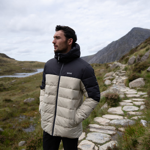 Man outdoors on stony trail wearing ALDTHORN puffer jacket in abbey stone with black shoulders and hood.