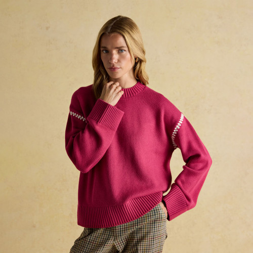 Woman wearing a relaxed-fit bright pink jumper with a chunky ribbed crew neck, cuffs, and hem. The sleeves feature a white zigzag stitch detail near the shoulders. Styled with checked trousers, the outfit is set against a soft beige background.