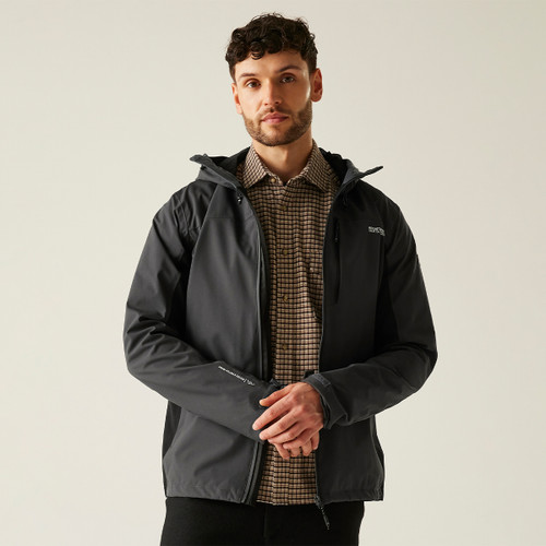 Man in a dark jacket over a plaid shirt stands against a plain background, exuding a relaxed and confident demeanour, with a neutral expression.