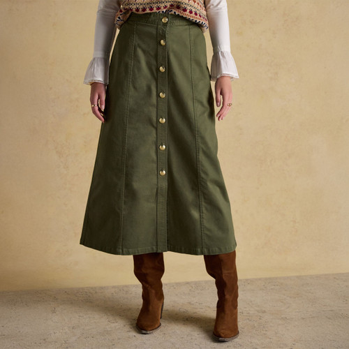 A woman wearing a long A-line olive green skirt with prominent front-facing gold buttons. The skirt has structured seam detailing and is paired with a cream blouse featuring flared cuffs and a patterned knit vest. She completes the outfit with knee-high brown suede boots, standing against a plain beige background.