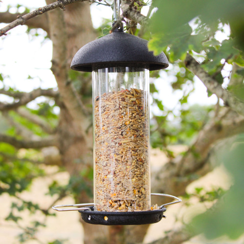 A cylindrical bird feeder filled with mixed seeds hangs from a tree branch. The background is blurred, showing leaves and tree bark in a natural setting.