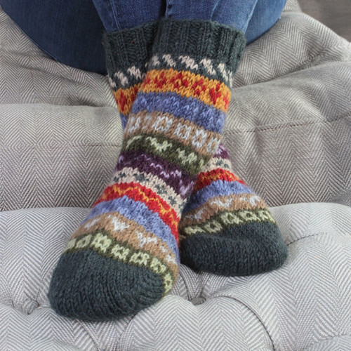 Cosy multicoloured knit socks with intricate patterns are worn by a person sitting cross-legged on a textured grey cushion, conveying warmth and comfort.