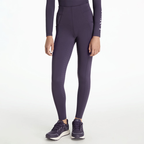 A person wearing dark purple LeMieux riding tights paired with matching long-sleeve top, shown from the front. The tights feature subtle silicone grip patterns along the inner legs, deep side pockets, and a smooth high waistband. The model is wearing purple trainers with white soles.