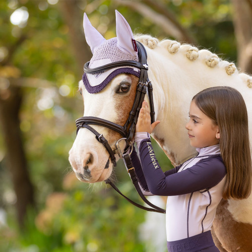 Young girl in a lilac and navy long-sleeved riding top gently touching the head of a cream and brown horse with braided mane, wearing a lilac and purple LeMieux fly hood and black bridle, in an outdoor setting with blurred greenery in the background.