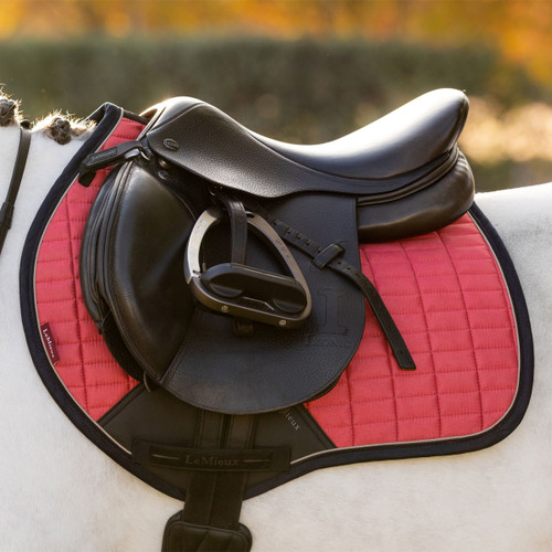 Close-up of a black leather saddle with black stirrups on a white quilted saddle pad featuring a bright red square-quilted design and black trim, secured on a white horse.