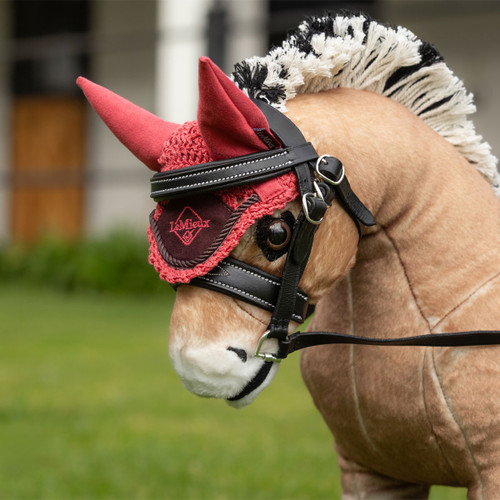A close-up of a tan and white plush horse toy wearing a red crocheted ear fly hood with matching red fabric ears and a dark red patch embroidered with the LeMieux logo, secured under a black leather bridle with silver buckles, against a blurred grassy background.