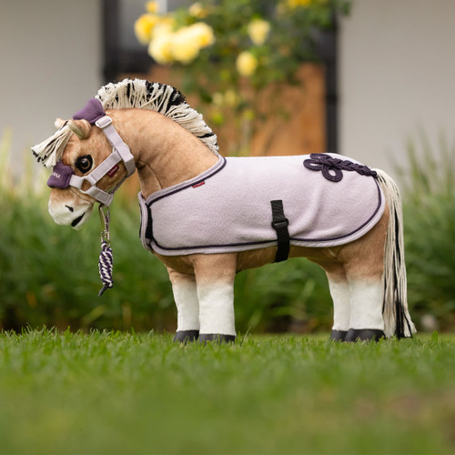 A tan plush toy horse stands on grass, wearing a light purple fleece rug with black trim and a black belly strap. It has a purple and white halter with a matching lead rope, and a black-and-white yarn mane.