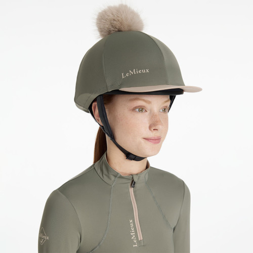 Rosemary green riding helmet silk with beige peak trim and faux fur pom-pom, paired with a matching quarter-zip base layer.