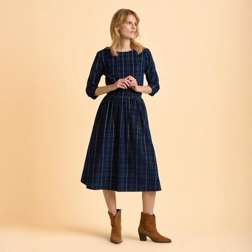 A woman in a blue plaid dress stands with hands clasped, wearing brown ankle boots. She gazes to the side against a light peach background.
