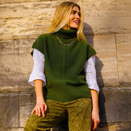 Woman in green turtleneck vest and patterned pants leans against a stone wall, smiling in the sunlight. Her pose is relaxed and content.
