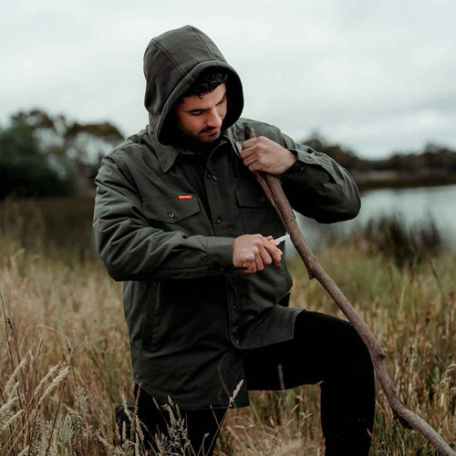 Man in a green jacket with a hood whittles a stick with a knife, kneeling in a grassy field by water. The scene feels focused and serene.