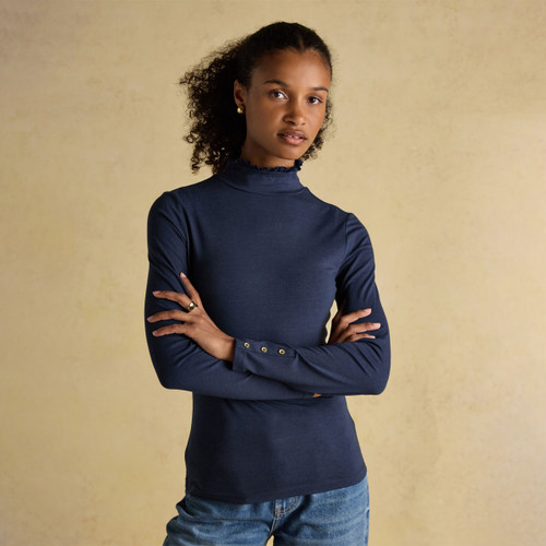 Woman wearing a navy frill-neck long-sleeve top with gold button cuff details, paired with blue jeans against a neutral backdrop.