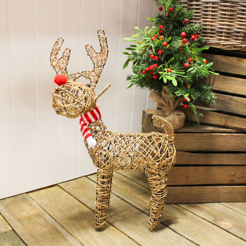 Wicker reindeer Christmas decoration with a red nose and striped scarf, standing on a wooden floor beside a small decorated artificial Christmas tree with red berries.